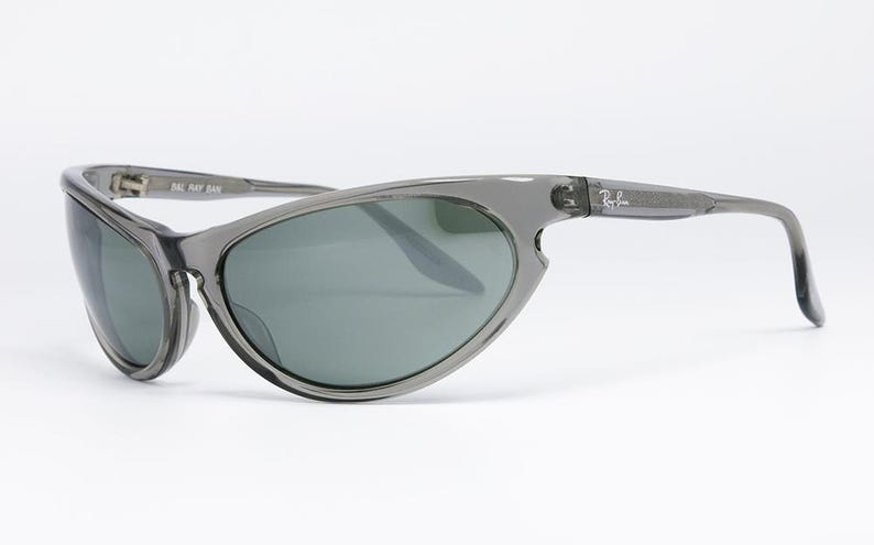 May include: Gray Ray-Ban sunglasses with a wraparound design. The lenses are a dark green color, and the frames are a translucent gray. The Ray-Ban logo is visible on the temple.