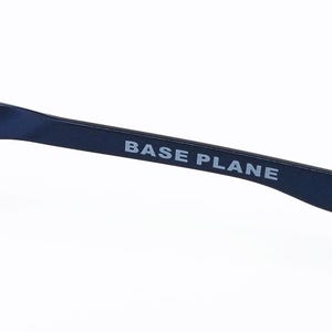 K&ouml;nnte beinhalten: Nahaufnahme eines marineblauen Brillenb&uuml;gels mit dem Text "BASE PLANE" in Wei&szlig;. Der B&uuml;gel hat ein schlankes, modernes Design und eine wei&szlig;e Spitze.