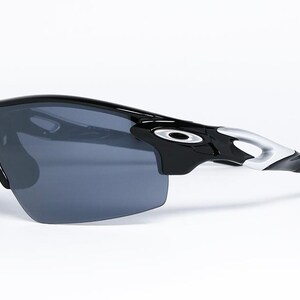 May include: Black sports sunglasses with a wraparound design. The sunglasses have a glossy black frame with silver accents on the temples and dark gray lenses. The Oakley logo is visible on the temples.