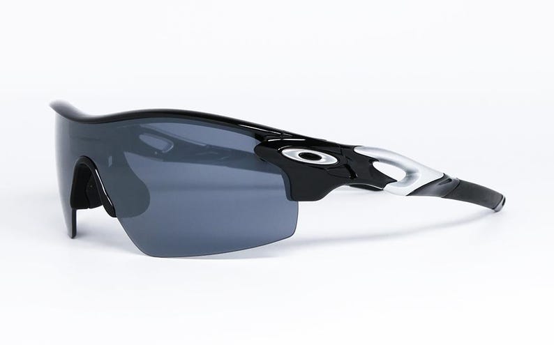 May include: Black sports sunglasses with a wraparound design. The sunglasses have a glossy black frame with silver accents on the temples and dark gray lenses. The Oakley logo is visible on the temples.