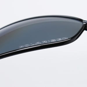 Puede incluir: Primer plano de gafas de sol negras con lentes grises. La palabra "POLARIZED" est&aacute; impresa en blanco en la lente. Las gafas de sol tienen un dise&ntilde;o elegante y moderno.