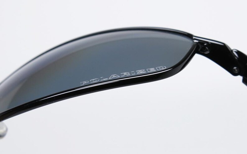 Puede incluir: Primer plano de gafas de sol negras con lentes grises. La palabra "POLARIZED" est&aacute; impresa en blanco en la lente. Las gafas de sol tienen un dise&ntilde;o elegante y moderno.