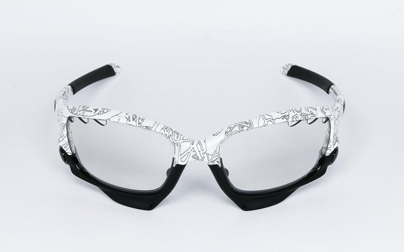 May include: White and black sports sunglasses with a unique design. The frames are white with a black pattern, and the arms and lower frame are black. The lenses are clear, and the overall style is modern and sporty.