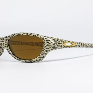 May include: Leopard print sunglasses with brown lenses. The frames feature a tan and black spotted pattern. The Oakley logo is visible on the temples. These sunglasses are a stylish accessory for eye protection.