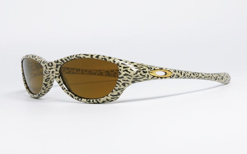 May include: Leopard print sunglasses with brown lenses. The frames feature a tan and black spotted pattern. The Oakley logo is visible on the temples. These sunglasses are a stylish accessory for eye protection.