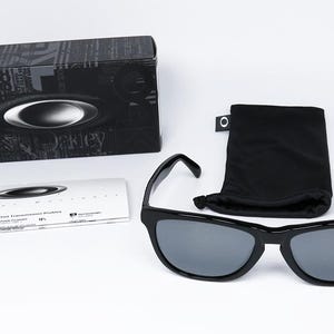 May include: Black sunglasses with gray lenses, a black case, a black drawstring pouch, and a white card. The sunglasses have a red logo on the bottom right lens. The case has the brand name and logo.