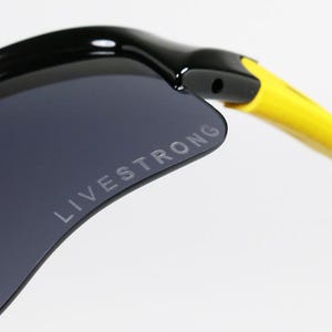 K&ouml;nnte beinhalten: Nahaufnahme einer schwarzen Sonnenbrille mit einer dunkelgrauen Linse. Der Text "LIVESTRONG" ist auf die Linse gedruckt. Der B&uuml;gel der Sonnenbrille ist gelb und schwarz.