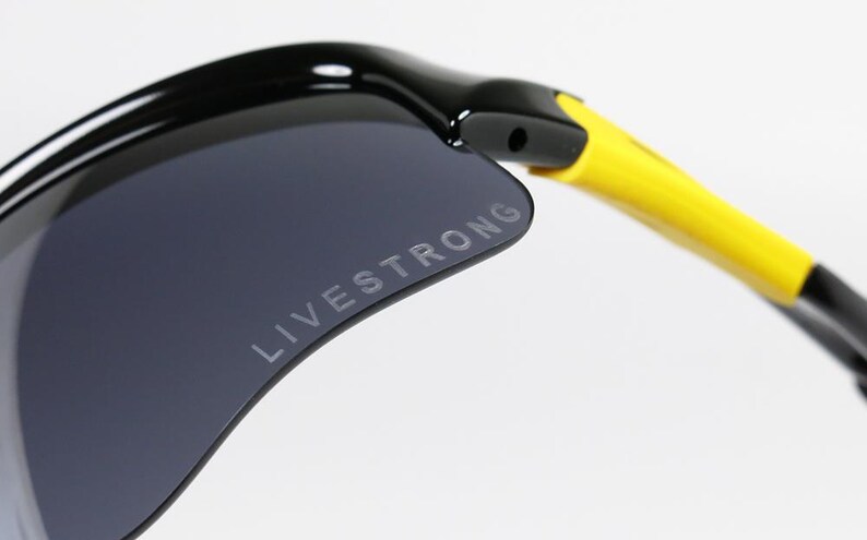 K&ouml;nnte beinhalten: Nahaufnahme einer schwarzen Sonnenbrille mit einer dunkelgrauen Linse. Der Text "LIVESTRONG" ist auf die Linse gedruckt. Der B&uuml;gel der Sonnenbrille ist gelb und schwarz.