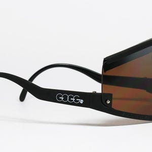 Op de afbeelding: Zwarte zonnebril met een wrap-around design en bruin getinte glazen. Het woord "GOGGs" is in het wit op de poot gedrukt. Het montuur heeft een matte zwarte afwerking.
