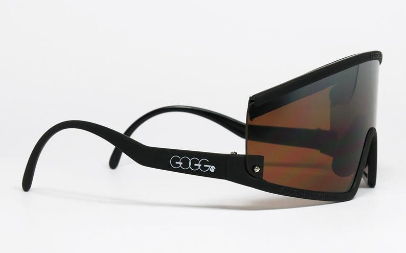 Op de afbeelding: Zwarte zonnebril met een wrap-around design en bruin getinte glazen. Het woord "GOGGs" is in het wit op de poot gedrukt. Het montuur heeft een matte zwarte afwerking.