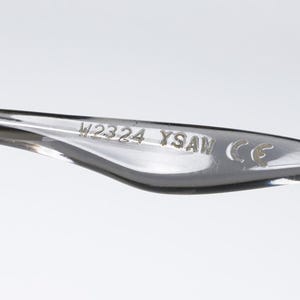 May include: Close-up of a clear eyeglass arm. The text "W2324 YSAM CE" is etched into the arm. The arm is curved and has a smooth, glossy finish. The background is a solid, light gray.