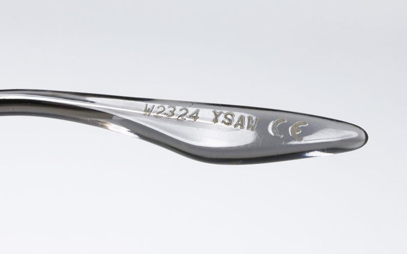 May include: Close-up of a clear eyeglass arm. The text "W2324 YSAM CE" is etched into the arm. The arm is curved and has a smooth, glossy finish. The background is a solid, light gray.