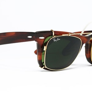 May include: Brown tortoiseshell sunglasses with green lenses and gold-colored frames. The sunglasses have a clip-on design and the Ray-Ban logo is visible on the lens. The glasses are set against a white background.