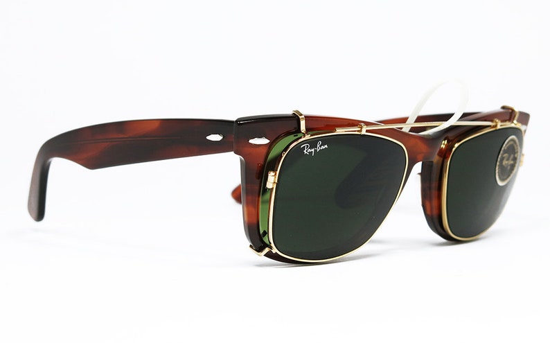 May include: Brown tortoiseshell sunglasses with green lenses and gold-colored frames. The sunglasses have a clip-on design and the Ray-Ban logo is visible on the lens. The glasses are set against a white background.