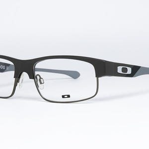 May include: Black and gray Oakley Yarddog II eyeglasses. The frames are a matte black with gray temples and the Oakley logo. The lenses are clear. The glasses are set against a white background.