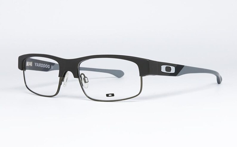 May include: Black and gray Oakley Yarddog II eyeglasses. The frames are a matte black with gray temples and the Oakley logo. The lenses are clear. The glasses are set against a white background.