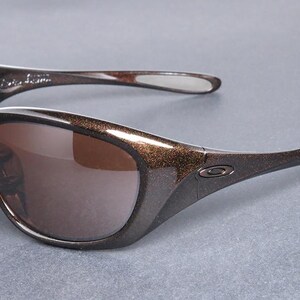 K&ouml;nnte beinhalten: Braune und schwarze Sonnenbrille mit geschwungenem Design. Die Gl&auml;ser sind braun und die Rahmen sind dunkelbraun mit einer gesprenkelten, glitzernden Textur. Die B&uuml;gel sind schwarz mit einer wei&szlig;en Spitze.