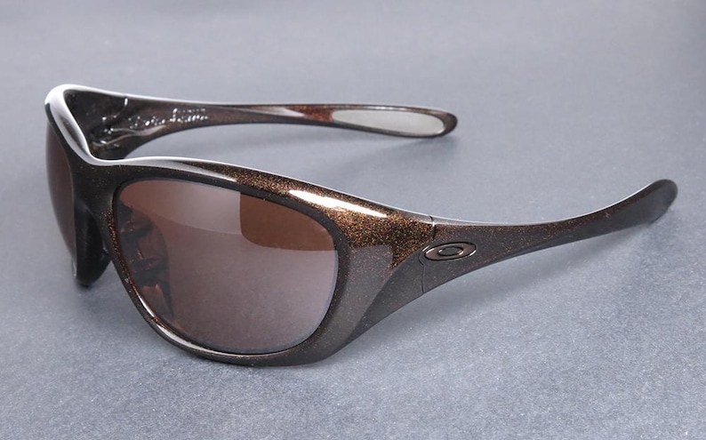 K&ouml;nnte beinhalten: Braune und schwarze Sonnenbrille mit geschwungenem Design. Die Gl&auml;ser sind braun und die Rahmen sind dunkelbraun mit einer gesprenkelten, glitzernden Textur. Die B&uuml;gel sind schwarz mit einer wei&szlig;en Spitze.
