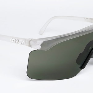 Puede incluir: Gafas de sol Oakley blancas transl&uacute;cidas con lentes verde oscuro. Las gafas tienen un dise&ntilde;o angular y futurista.