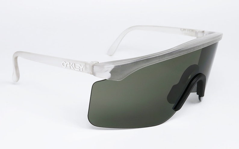 Puede incluir: Gafas de sol Oakley blancas transl&uacute;cidas con lentes verde oscuro. Las gafas tienen un dise&ntilde;o angular y futurista.