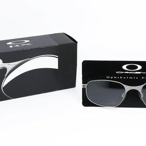 May include: A pair of silver-toned Oakley RX ophthalmic frames with dark gray lenses. The frames are displayed on a black background with the Oakley logo and the text "Ophthalmic Frame".