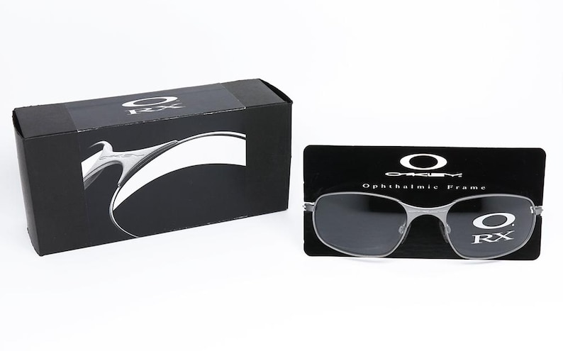 May include: A pair of silver-toned Oakley RX ophthalmic frames with dark gray lenses. The frames are displayed on a black background with the Oakley logo and the text "Ophthalmic Frame".