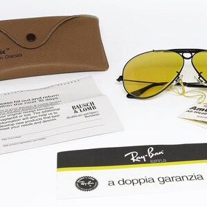 Puede incluir: Gafas de sol Ray-Ban AmberMatic con lentes amarillas y montura negra, junto con un estuche de cuero marr&oacute;n y documentos. El estuche y las gafas lucen los logotipos "Ray-Ban" y "AmberMatic".