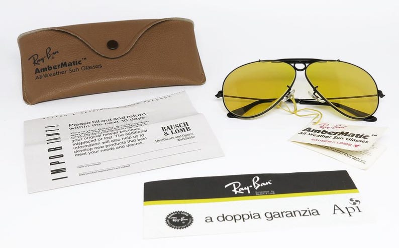Puede incluir: Gafas de sol Ray-Ban AmberMatic con lentes amarillas y montura negra, junto con un estuche de cuero marr&oacute;n y documentos. El estuche y las gafas lucen los logotipos "Ray-Ban" y "AmberMatic".