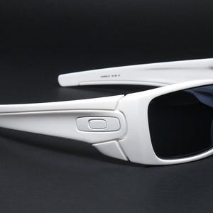 May include: White sunglasses with dark lenses. The sunglasses have a rectangular frame with a curved design. The arms of the sunglasses are thick and have a slight curve. The lenses are dark, and the frame is white.