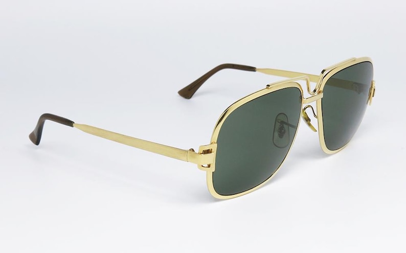 Puede incluir: Gafas de sol doradas con lentes verde oscuro. Las gafas de sol tienen un dise&ntilde;o cl&aacute;sico de aviador con doble puente y forma rectangular. Las patillas son finas y doradas, con puntas marrones. Las lentes son de color verde oscuro.