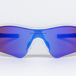 Pu&ograve; includere: Occhiali da sole con montatura bianca e lenti a specchio blu e rosse. Gli occhiali hanno un design curvo e il marchio "OAKLEY" &egrave; stampato sulla lente. Ideali per lo sport e le attivit&agrave; all'aperto.