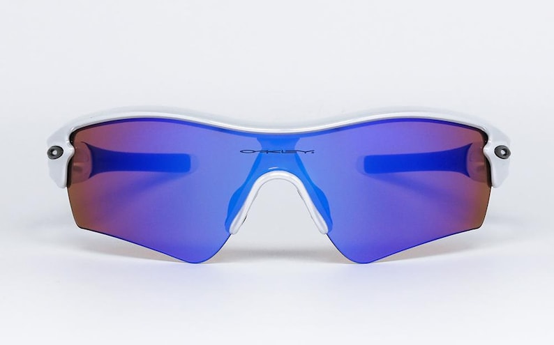 Pu&ograve; includere: Occhiali da sole con montatura bianca e lenti a specchio blu e rosse. Gli occhiali hanno un design curvo e il marchio "OAKLEY" &egrave; stampato sulla lente. Ideali per lo sport e le attivit&agrave; all'aperto.