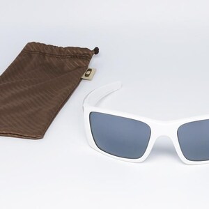 May include: White sunglasses with grey lenses and a brown drawstring pouch. The sunglasses have a rectangular frame and a modern design. The pouch is made of a soft, textured material.