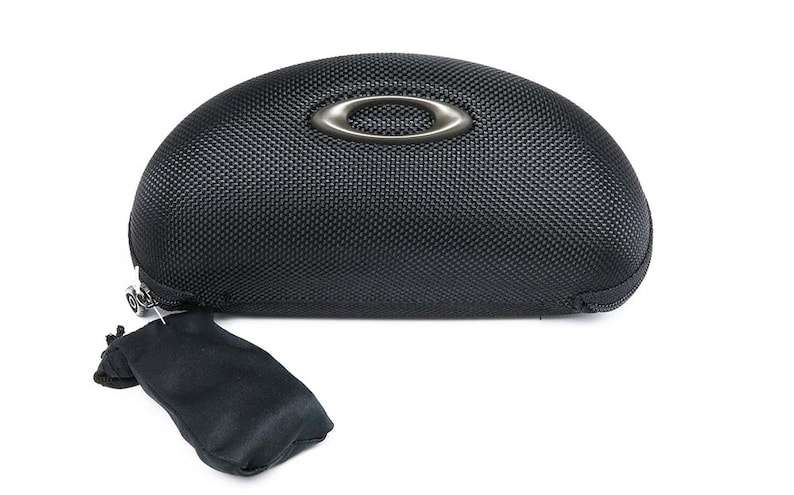 May include: Black, textured eyewear case with a silver-tone Oakley logo. The case has a zipper closure and includes a small black pouch. The case is designed to protect eyeglasses or sunglasses.