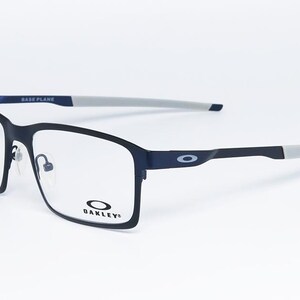 K&ouml;nnte beinhalten: Rechteckige Brille in Schwarz und Marineblau mit klaren Gl&auml;sern. Die Fassungen haben eine matte Oberfl&auml;che mit grauen B&uuml;gelenden. Das Oakley-Logo ist auf dem Rahmen und dem B&uuml;gel sichtbar. Der Text "BASE PLANE" ist auf der Oberseite des Rahmens aufgedruckt.