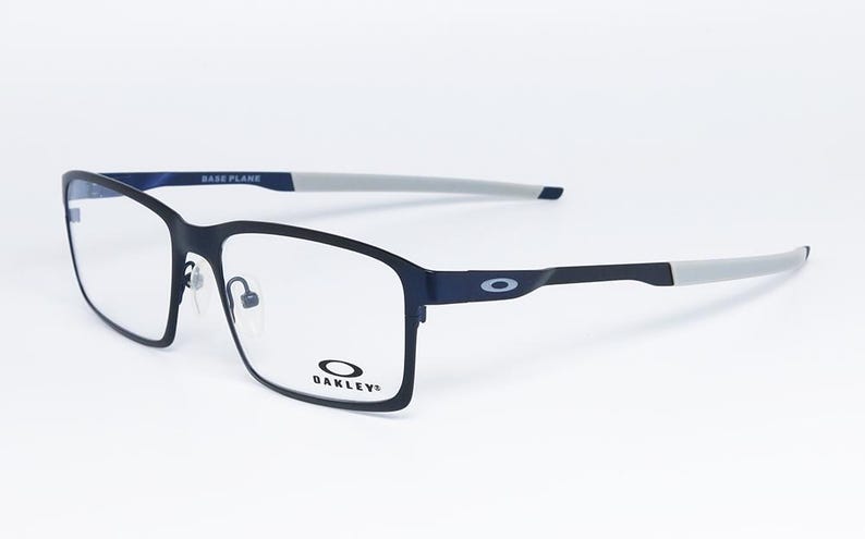 K&ouml;nnte beinhalten: Rechteckige Brille in Schwarz und Marineblau mit klaren Gl&auml;sern. Die Fassungen haben eine matte Oberfl&auml;che mit grauen B&uuml;gelenden. Das Oakley-Logo ist auf dem Rahmen und dem B&uuml;gel sichtbar. Der Text "BASE PLANE" ist auf der Oberseite des Rahmens aufgedruckt.