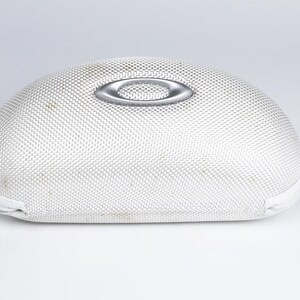 May include: White, oval-shaped Oakley sunglasses case with a silver logo on top. The case has a textured surface and a zipper closure. The case is designed to protect eyewear.