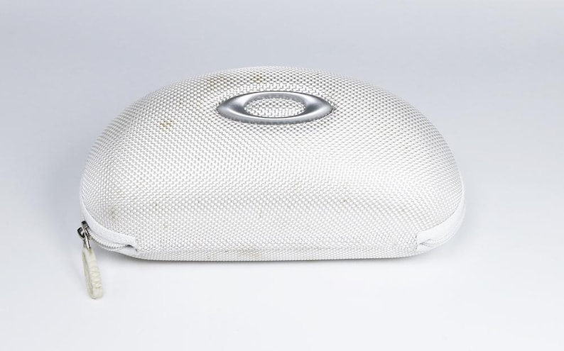 May include: White, oval-shaped Oakley sunglasses case with a silver logo on top. The case has a textured surface and a zipper closure. The case is designed to protect eyewear.