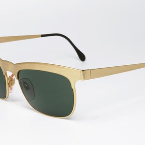 Puede incluir: Gafas de sol doradas con lentes verde oscuro. Las monturas tienen forma rectangular con acabado mate. Las patillas son finas y se extienden hasta terminales negros curvados. Un accesorio de gafas cl&aacute;sico.