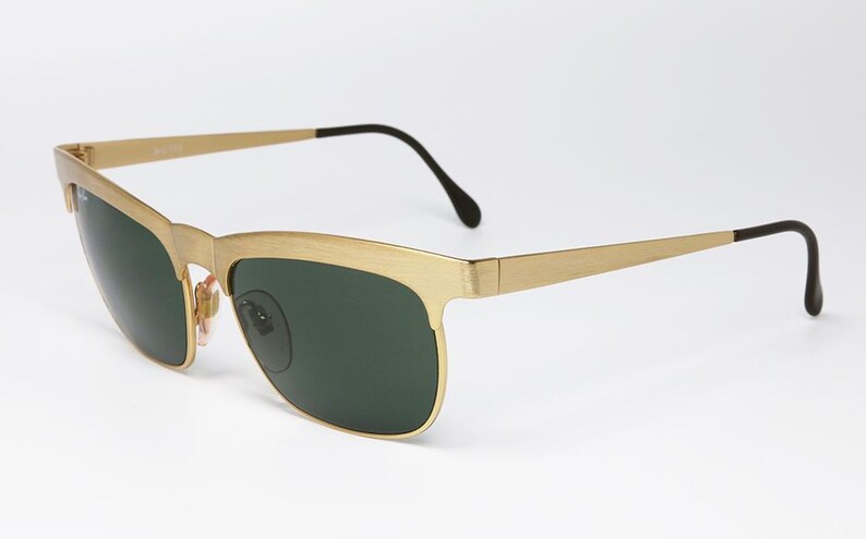 Puede incluir: Gafas de sol doradas con lentes verde oscuro. Las monturas tienen forma rectangular con acabado mate. Las patillas son finas y se extienden hasta terminales negros curvados. Un accesorio de gafas cl&aacute;sico.