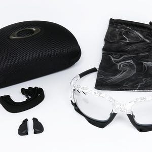May include: An assortment of Oakley eyewear accessories, including a black hard case, a black drawstring bag, a pair of sunglasses with a white patterned frame, and extra nose pads. The sunglasses have clear lenses.