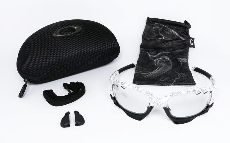 May include: An assortment of Oakley eyewear accessories, including a black hard case, a black drawstring bag, a pair of sunglasses with a white patterned frame, and extra nose pads. The sunglasses have clear lenses.