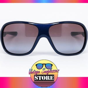 May include: Dark blue sunglasses with a wide frame and gradient lenses. The sunglasses have a rounded shape and a thick, sturdy frame. The lenses transition from a darker shade at the top to a lighter shade at the bottom. The logo of the Vintage Sunglasses Store is at the bottom.