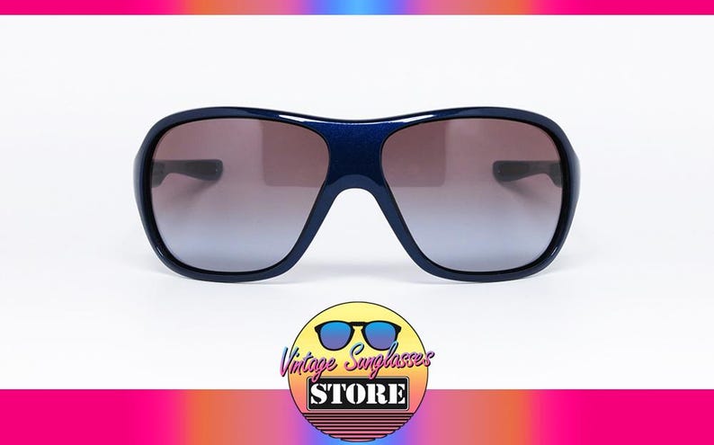 May include: Dark blue sunglasses with a wide frame and gradient lenses. The sunglasses have a rounded shape and a thick, sturdy frame. The lenses transition from a darker shade at the top to a lighter shade at the bottom. The logo of the Vintage Sunglasses Store is at the bottom.