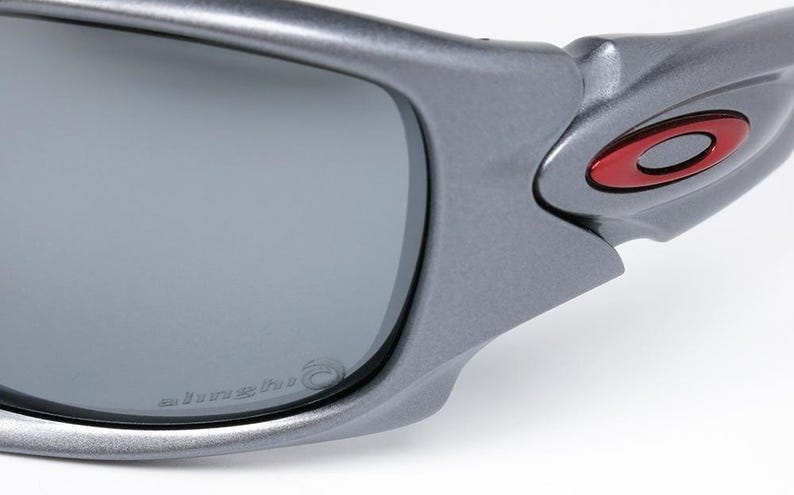 May include: Close-up of a pair of gray sunglasses with a red and black logo on the temple. The lenses are gray and have a subtle logo etched on the bottom corner. The frame is a matte gray.