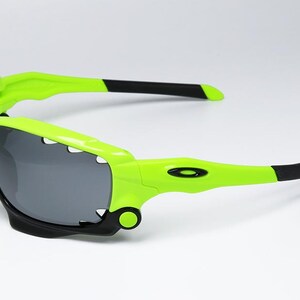 May include: Neon green and black sports sunglasses with gray lenses. The sunglasses have a distinctive, angular design with a bright green frame and black accents on the arms. The lenses are a dark gray color.