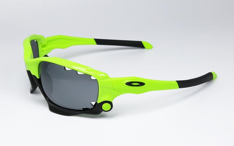 May include: Neon green and black sports sunglasses with gray lenses. The sunglasses have a distinctive, angular design with a bright green frame and black accents on the arms. The lenses are a dark gray color.
