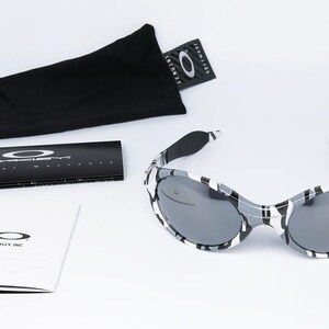 May include: Sunglasses with a black and white camouflage pattern. The sunglasses have dark lenses and are accompanied by a black carrying case, a cleaning cloth, and product literature. The Oakley logo is visible on the glasses and packaging.