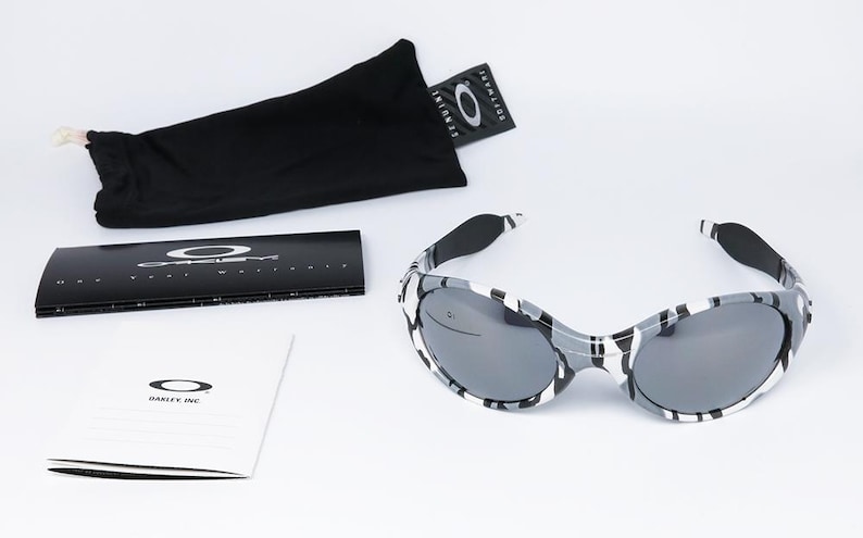 May include: Sunglasses with a black and white camouflage pattern. The sunglasses have dark lenses and are accompanied by a black carrying case, a cleaning cloth, and product literature. The Oakley logo is visible on the glasses and packaging.