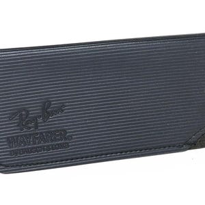 May include: A dark blue Ray-Ban Wayfarer glasses case. The case has a textured, ribbed surface and a black suede corner. The Ray-Ban logo and "WAYFARER BY BAUSCH & LOMB" are embossed on the front.